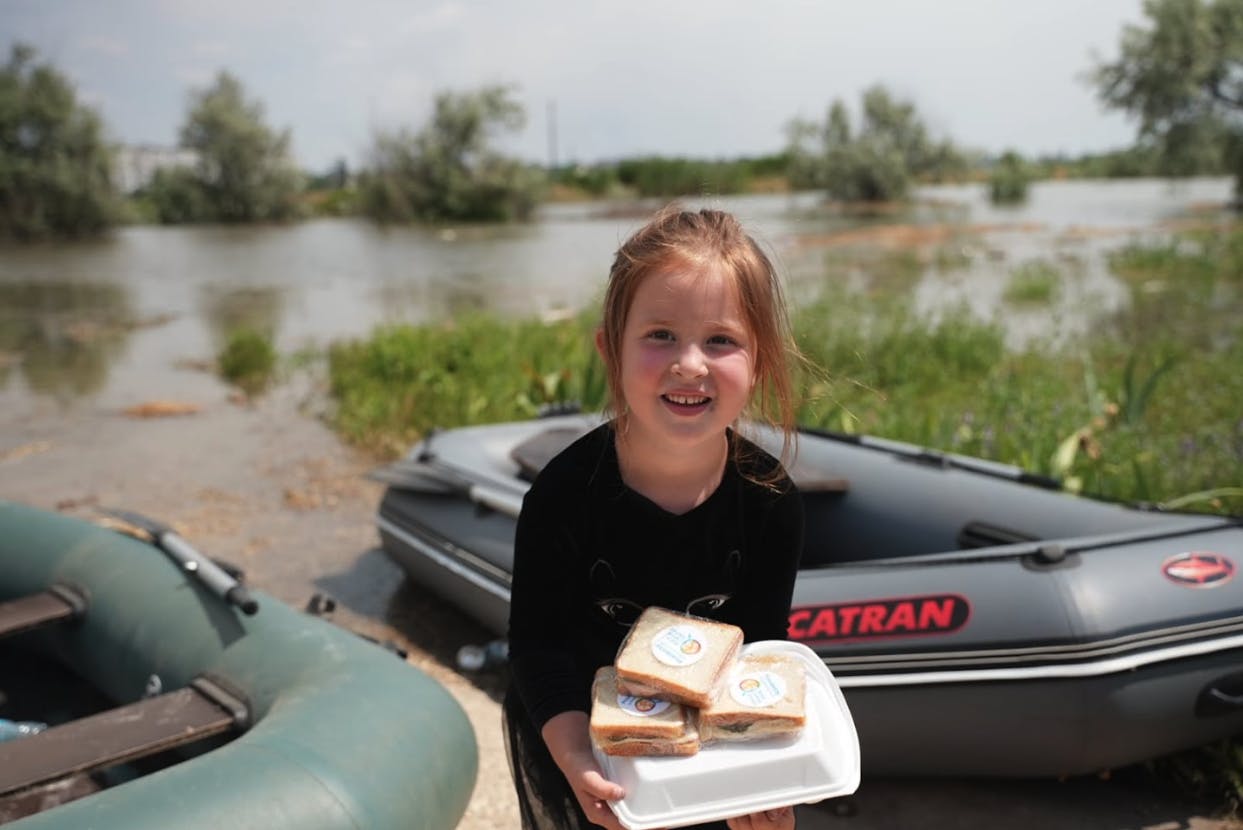 A young girl holds a hot meal and three sandwiches with flooding and two boats visible behind her. 